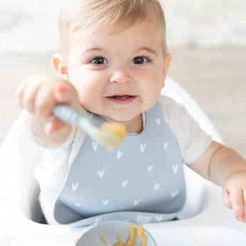 A baby in a high chair uses Bumkins Silicone Bib: Hearts, featuring a light blue design with white hearts and a convenient pocket.