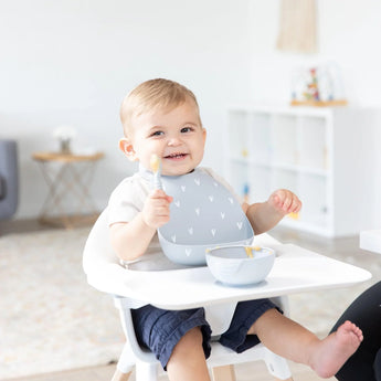 A happy baby in a high chair wears the Bumkins Silicone Bib: Hearts with catch-all pocket, holding a spoon by their food bowl. Bright room.