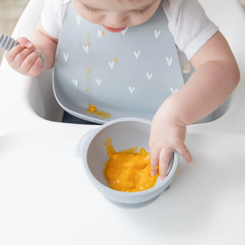 A baby in a high chair, wearing a Bumkins Silicone Bib: Hearts with a catch-all pocket, plays with orange puree and holds a spoon.