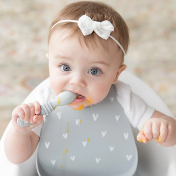 The baby with a white bow enjoys using a spoon and the Bumkins Silicone Bib: Hearts, featuring heart patterns and a catch-all pocket.