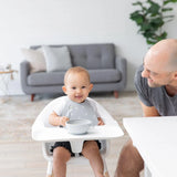 A baby in a high chair smiles with a spoon, wearing the Bumkins Silicone Bib: Hearts. An adult leans over; a gray sofa and plant are behind.