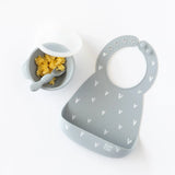 Bumkins Silicone Bib: Hearts in gray with a catch-all pocket next to a suction bowl of eggs on a white backdrop.