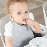 A curly-haired toddler with a messy face eats with a spoon, wearing a Bumkins Silicone Bib: Hearts, featuring a catch-all pocket.