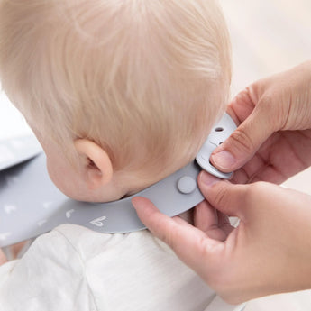 Adult hands fasten a Bumkins Silicone Bib: Hearts on a babys neck over a white shirt.