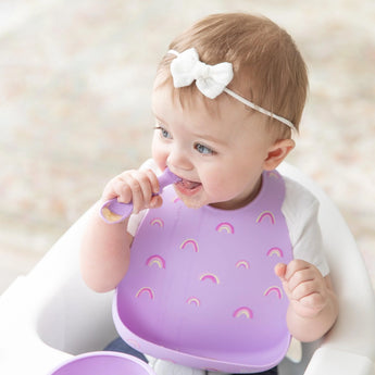 A baby in a bow headband uses a spoon, wearing Bumkins Rainbows silicone bib in the high chair to manage mealtime mess.