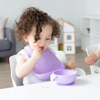 A toddler with curly hair enjoys a meal from a purple bowl, wearing Bumkins Silicone Bib: Rainbows, seated safely indoors.