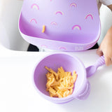 A child in a Bumkins Silicone Bib: Rainbows enjoys mac and cheese from a matching purple bowl, keeping mealtime mess-free.