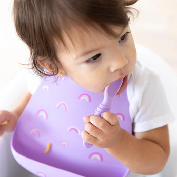 A toddler enjoys mealtime with a Bumkins Rainbows silicone bib and purple spoon, safely seated in their high chair.