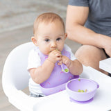 A baby in a highchair uses a Bumkins Silicone Bib: Rainbows, eating with a spoon from a purple bowl, for mess-free mealtime.