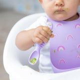 A baby in a high chair, wearing Bumkins Silicone Bib: Rainbows, skillfully maneuvers a purple spoon amid the adorable mealtime chaos.