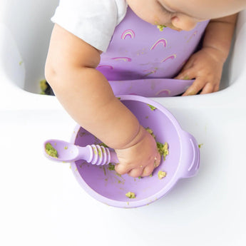 A baby in a Bumkins Silicone Bib: Rainbows joyfully creates a mealtime mess with avocado, safely seated in the high chair.