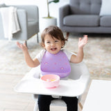 A toddler in a highchair, wearing a Bumkins Silicone Bib: Rainbows, smiles with raised arms at mealtime alongside a bowl and sofa.