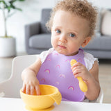 A curly-haired baby in a Bumkins Rainbows silicone bib holds a spoon, ready to eat by a yellow bowl, with a couch in the background.