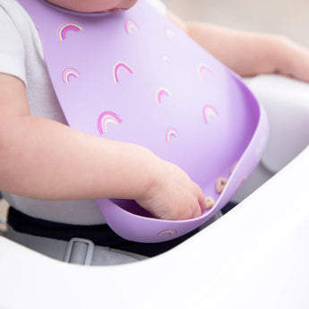 A baby in a high chair wears a purple Bumkins Silicone Bib with Rainbows, reaching into the pocket filled with snacks.