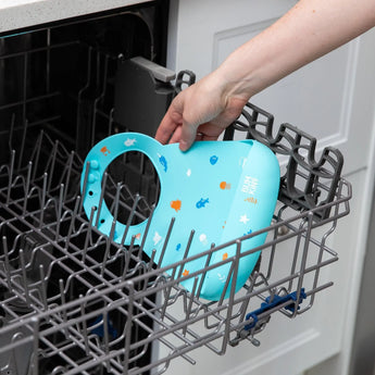 A hand places the Bumkins Silicone Bib: Ocean Life into the dishwashers top rack.