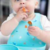 A baby with a Silicone Bib: Ocean Life by Bumkins tackles mealtime, enjoying orange food with a spoon and some on their fingers.