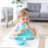 A toddler in a highchair, wearing Bumkins Silicone Bib: Ocean Life, smiles with spoon in hand amidst the mealtime mess. Cozy room behind.