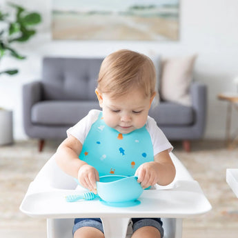 A baby enjoys mealtime in a bright room, wearing a Bumkins Silicone Bib: Ocean Life and playing with a blue bowl and spoon.