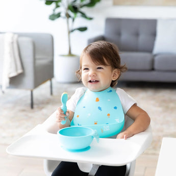 A toddler in a high chair, wearing a Bumkins Silicone Bib: Ocean Life, smiles with a spoon and blue bowl amid mealtime fun.