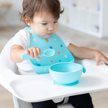 A toddler in a Bumkins Silicone Bib: Ocean Life holds a spoon and eyes the bowl of food on the high chair tray.