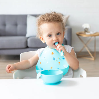 A toddler with curly hair in a highchair wears a Bumkins Silicone Bib: Ocean Life, holding a spoon, blue bowl in front, sofa behind.