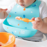 A baby sporting Bumkins Ocean Life silicone bib uses an orange spoon and bowl, diving into mealtime fun.