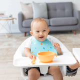 A baby in a highchair smiles during mealtime, wearing a Bumkins Silicone Bib: Ocean Life, eating from an orange bowl. Sofa and table behind.