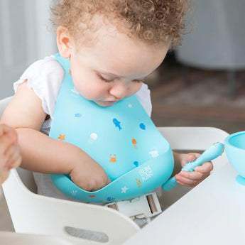 Toddler in high chair wearing a Bumkins Ocean Life Silicone Bib, holding a spoon, eagerly reaching into a bowl during mealtime.