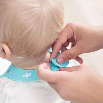 Hands fastening a Bumkins Silicone Bib: Ocean Life in blue, around a babys neck, ready to tackle any mealtime mess.