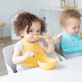 Toddlers enjoy a mealtime mess using Bumkins Silicone Bibs: Camp Gear; one uses a spoon with a yellow bowl, the other sips from a cup.