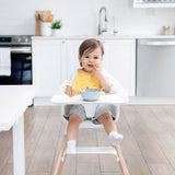 A baby in a high chair wears a Bumkins Silicone Bib: Camp Gear, smiling while holding food in a modern kitchen.