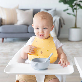 A smiling toddler in a high chair wears a Bumkins Silicone Bib: Camp Gear, embracing the mealtime mess with a spoon.