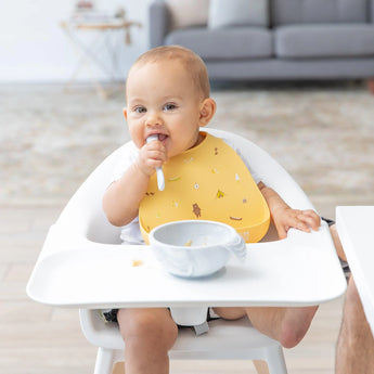 A baby in a high chair wearing a Bumkins Camp Gear Silicone Bib eagerly tackles mealtime, enjoying food with a spoon and bowl.