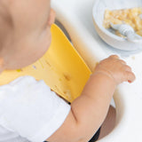 A baby delights in mess at meals, wearing a Bumkins Camp Gear yellow silicone bib, with mashed food and spoon nearby.