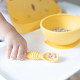 A child grips a yellow spoon with food beside a matching bowl, ready to tackle mealtime mess in a Bumkins Silicone Bib: Camp Gear.