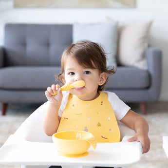 A toddler in a Bumkins Silicone Bib: Camp Gear sits in a high chair with a yellow spoon and bowl; a gray sofa is seen behind them.