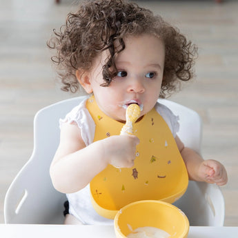 A curly-haired toddler in a high chair with a Bumkins Camp Gear silicone bib, holding a spoon and enjoying mealtime with a yellow bowl.