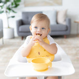 A toddler with a Bumkins Silicone Bib: Camp Gear sits in a high chair, holding food near a yellow bowl. A gray sofa and plant are behind.