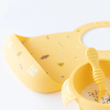 Bumkins Silicone Bib: Camp Gear, paired with a cereal bowl and spoon, keeps mealtime mess away on a white background.