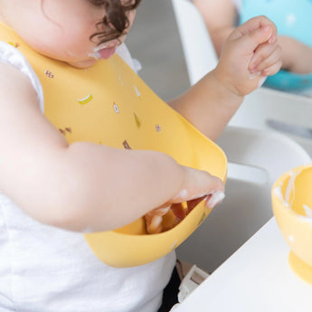 A toddler in a yellow Bumkins Silicone Bib: Camp Gear messily enjoys creamy food, smearing it delightfully on their face and bib.