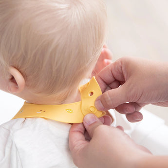 Hands fasten a Bumkins Silicone Bib: Camp Gear around a babys neck, ready to tackle mealtime mess from the back view.