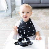 A smiling toddler in a high chair wears a Bumkins Silicone Bib: Mickey Mouse Faces, holding a spoon by a food-filled bowl.