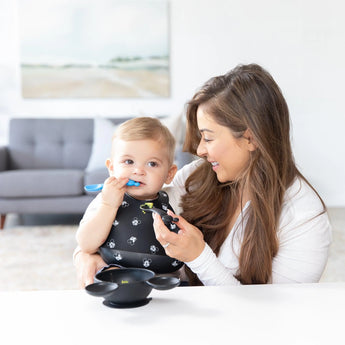 A woman smiles at a baby in a Bumkins Mickey Mouse Faces Silicone Bib, feeding them with a blue spoon in a bright, art-filled room.