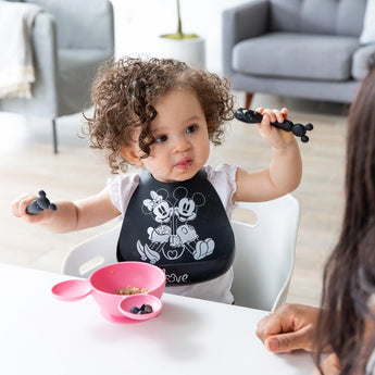 A curly-haired toddler in a high chair wears a Bumkins Mickey + Minnie Mouse silicone bib, surrounded by toys and food.