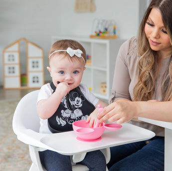 A baby in a high chair with a pink bowl wears Bumkins Silicone Bib: Mickey + Minnie Mouse, while an adult assists beside him.