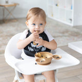 A toddler in a highchair, wearing a Bumkins Silicone Bib: Mickey + Minnie Mouse, eagerly munches snacks with food in both hands.