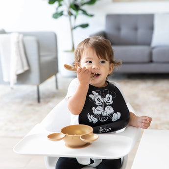 A toddler in a Bumkins Silicone Bib: Mickey + Minnie Mouse eats from a bowl in a high chair, with a gray couch behind.