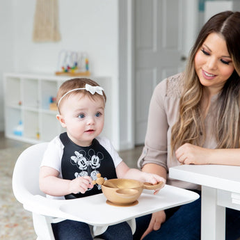 A baby in a high chair with a Bumkins Mickey + Minnie Mouse bib and bowl is watched by a smiling woman beside them.