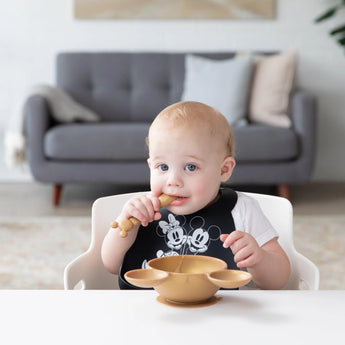 A baby in a high chair wears a Bumkins Silicone Bib: Mickey + Minnie Mouse, holding a spoon near a bowl; a gray sofa is in the background.