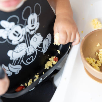 A child enjoys scrambled eggs in a bowl, wearing a Bumkins Silicone Bib: Mickey + Minnie Mouse, and drops food on the table.